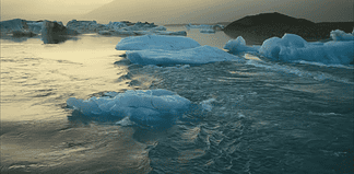 Icebergs floating in a calm glacial lagoon at sunset with distant mountains in the background, reflecting on the water.