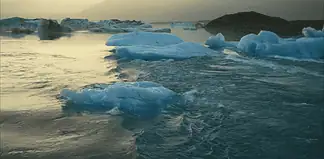 Icebergs floating in a calm glacial lagoon at sunset with distant mountains in the background, reflecting on the water.
