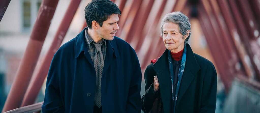 Man and elderly woman walk and chat on a red metal bridge, wearing coats and scarves.