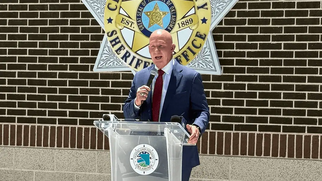 Weaver press conference Man in a navy suit and red tie speaks into a handheld microphone at a clear podium bearing a circular seal, with a sheriff’s office emblem on a brick wall behind him.