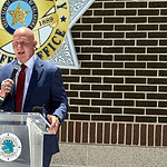 Man in a navy suit and red tie speaks into a handheld microphone at a clear podium bearing a circular seal, with a sheriff’s office emblem on a brick wall behind him.