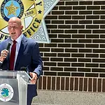 Weaver named new Cleveland Co. sheriff; no FBI investigation expected Man in a navy suit and red tie speaks into a handheld microphone at a clear podium bearing a circular seal, with a sheriff’s office emblem on a brick wall behind him.