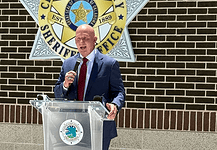 Man in a navy suit and red tie speaks into a handheld microphone at a clear podium bearing a circular seal, with a sheriff’s office emblem on a brick wall behind him.