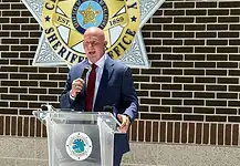 Weaver named new Cleveland Co. sheriff; no FBI investigation expected Man in a navy suit and red tie speaks into a handheld microphone at a clear podium bearing a circular seal, with a sheriff’s office emblem on a brick wall behind him.