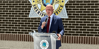 Man in a navy suit and red tie speaks into a handheld microphone at a clear podium bearing a circular seal, with a sheriff’s office emblem on a brick wall behind him.