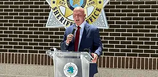 Man in a navy suit and red tie speaks into a handheld microphone at a clear podium bearing a circular seal, with a sheriff’s office emblem on a brick wall behind him.