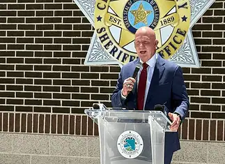 Weaver named new Cleveland Co. sheriff; no FBI investigation expected Man in a navy suit and red tie speaks into a handheld microphone at a clear podium bearing a circular seal, with a sheriff’s office emblem on a brick wall behind him.