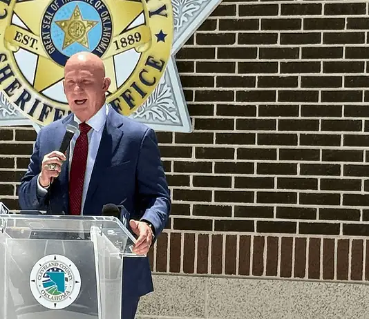 Weaver named new Cleveland Co. sheriff; no FBI investigation expected Man in a navy suit and red tie speaks into a handheld microphone at a clear podium bearing a circular seal, with a sheriff’s office emblem on a brick wall behind him.