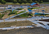 PRESS RELEASE: New north section of Wiley Post Park opens Aerial view of a modern playground with blue shade sails, slides, climbing structures, and winding concrete paths in a park setting.