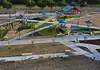 PRESS RELEASE: New north section of Wiley Post Park opens Aerial view of a modern playground with blue shade sails, slides, climbing structures, and winding concrete paths in a park setting.