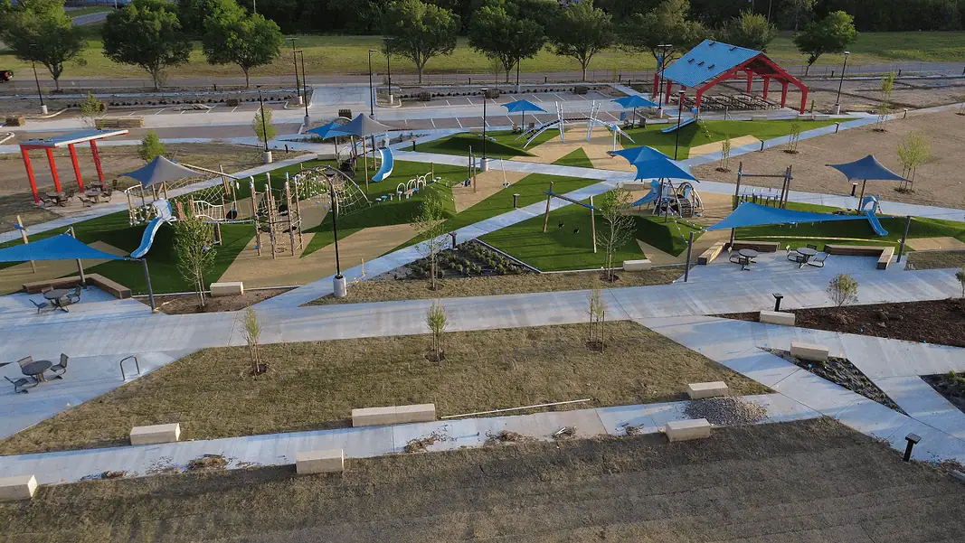 Wiley Post Park updates Aerial view of a modern playground with blue shade sails, slides, climbing structures, and winding concrete paths in a park setting.