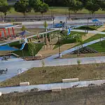 PRESS RELEASE: New north section of Wiley Post Park opens Aerial view of a modern playground with blue shade sails, slides, climbing structures, and winding concrete paths in a park setting.