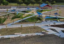 PRESS RELEASE: New north section of Wiley Post Park opens Aerial view of a modern playground with blue shade sails, slides, climbing structures, and winding concrete paths in a park setting.