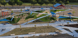 Aerial view of a modern playground with blue shade sails, slides, climbing structures, and winding concrete paths in a park setting.