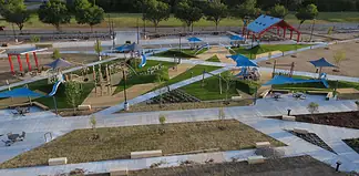Aerial view of a modern playground with blue shade sails, slides, climbing structures, and winding concrete paths in a park setting.