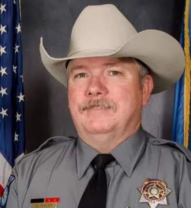 Portrait of a male sheriff deputy in a gray uniform with a black tie, wearing a white cowboy hat, standing in front of an American flag.