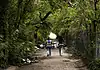 Housing voucher inspections slowing OKC homeless encampment closings Two workers in reflective safety vests walk down a tree-lined dirt path with litter on the ground and a chain-link fence to the right.