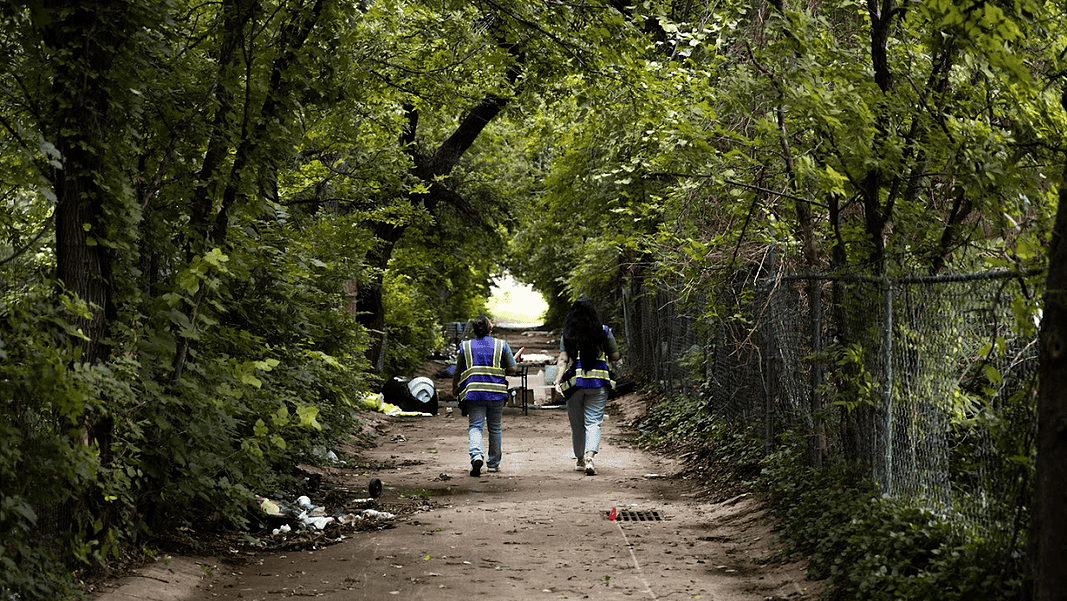 Two workers in reflective safety vests walk down a tree-lined dirt path with litter on the ground and a chain-link fence to the right.