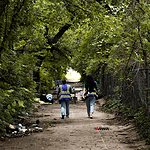 Two workers in reflective safety vests walk down a tree-lined dirt path with litter on the ground and a chain-link fence to the right.