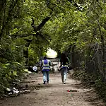 Housing voucher inspections slowing OKC homeless encampment closings Two workers in reflective safety vests walk down a tree-lined dirt path with litter on the ground and a chain-link fence to the right.