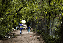 Two workers in reflective safety vests walk down a tree-lined dirt path with litter on the ground and a chain-link fence to the right.