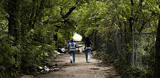 Two workers in reflective safety vests walk down a tree-lined dirt path with litter on the ground and a chain-link fence to the right.