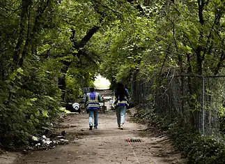 Housing voucher inspections slowing OKC homeless encampment closings Two workers in reflective safety vests walk down a tree-lined dirt path with litter on the ground and a chain-link fence to the right.