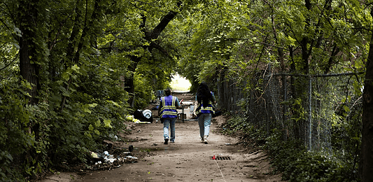 Two workers in reflective safety vests walk down a tree-lined dirt path with litter on the ground and a chain-link fence to the right.