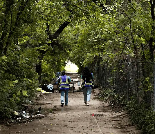 Housing voucher inspections slowing OKC homeless encampment closings Two workers in reflective safety vests walk down a tree-lined dirt path with litter on the ground and a chain-link fence to the right.