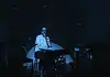 Creative covers, triumphant tributes drive upcoming concerts and releases Person seated at a keyboard in a dim, blue-lit room with green star projector specks on the walls and floor.