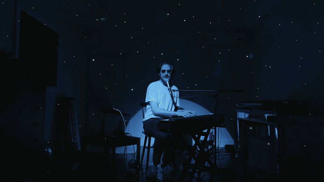 Bo Burnham 2 Person seated at a keyboard in a dim, blue-lit room with green star projector specks on the walls and floor.