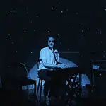 Creative covers, triumphant tributes drive upcoming concerts and releases Person seated at a keyboard in a dim, blue-lit room with green star projector specks on the walls and floor.