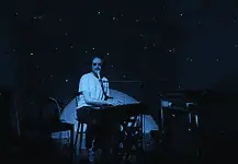 Creative covers, triumphant tributes drive upcoming concerts and releases Person seated at a keyboard in a dim, blue-lit room with green star projector specks on the walls and floor.