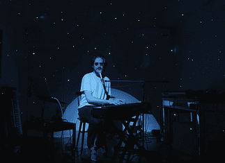 Creative covers, triumphant tributes drive upcoming concerts and releases Person seated at a keyboard in a dim, blue-lit room with green star projector specks on the walls and floor.
