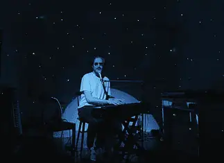 Creative covers, triumphant tributes drive upcoming concerts and releases Person seated at a keyboard in a dim, blue-lit room with green star projector specks on the walls and floor.