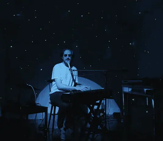 Creative covers, triumphant tributes drive upcoming concerts and releases Person seated at a keyboard in a dim, blue-lit room with green star projector specks on the walls and floor.