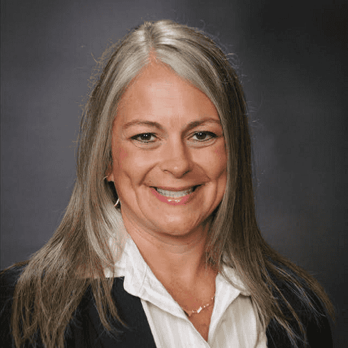 Professional headshot of a smiling woman with shoulder-length gray hair, wearing a white blouse and dark blazer against a neutral background.