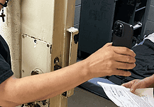 Person in a black shirt holds a smartphone to photograph printed papers being handed to them near a rusty metal door.