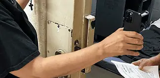 Person in a black shirt holds a smartphone to photograph printed papers being handed to them near a rusty metal door.
