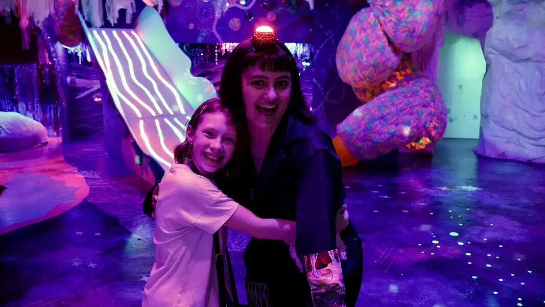 IMG_0146 Two people hug and smile in a neon-lit, immersive art space with colorful glowing sculptures behind them, purple lighting everywhere.