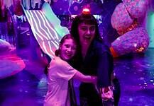 Two people hug and smile in a neon-lit, immersive art space with colorful glowing sculptures behind them, purple lighting everywhere.