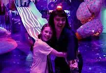 Factory Obscura’s RECORD+PLAY adds new layers of interaction to Mix-Tape Two people hug and smile in a neon-lit, immersive art space with colorful glowing sculptures behind them, purple lighting everywhere.