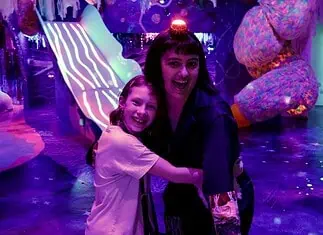 Factory Obscura’s RECORD+PLAY adds new layers of interaction to Mix-Tape Two people hug and smile in a neon-lit, immersive art space with colorful glowing sculptures behind them, purple lighting everywhere.