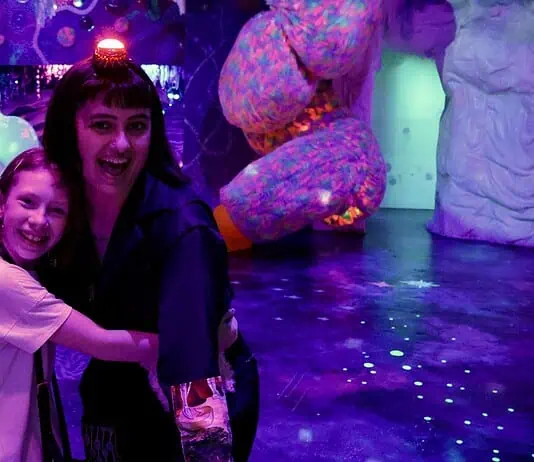 Factory Obscura’s RECORD+PLAY adds new layers of interaction to Mix-Tape Two people hug and smile in a neon-lit, immersive art space with colorful glowing sculptures behind them, purple lighting everywhere.