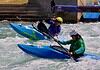 Two kayakers wearing helmets navigate whitewater rapids in blue kayaks, paddling aggressively through churning water.