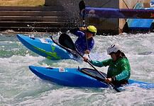 Two kayakers wearing helmets navigate whitewater rapids in blue kayaks, paddling aggressively through churning water.