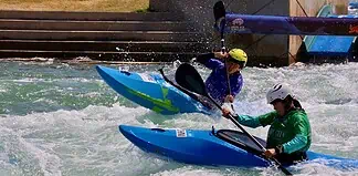 Two kayakers wearing helmets navigate whitewater rapids in blue kayaks, paddling aggressively through churning water.