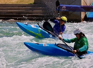 Riversport OKC scores big hosting World Championships ahead of Olympics Two kayakers wearing helmets navigate whitewater rapids in blue kayaks, paddling aggressively through churning water.