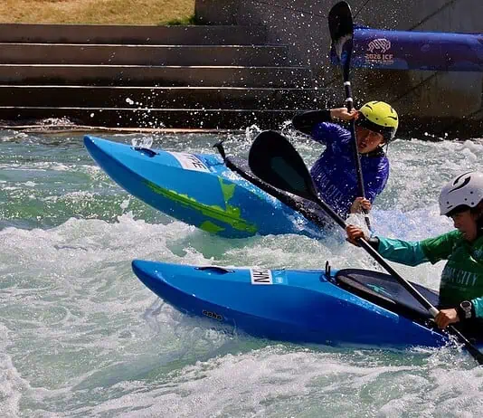 Riversport OKC scores big hosting World Championships ahead of Olympics Two kayakers wearing helmets navigate whitewater rapids in blue kayaks, paddling aggressively through churning water.