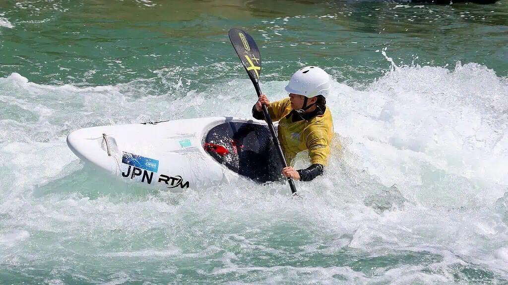 Kayaker wearing a white helmet and yellow wetsuit navigating whitewater in a white kayak with a double-bladed paddle on turbulent turquoise water.